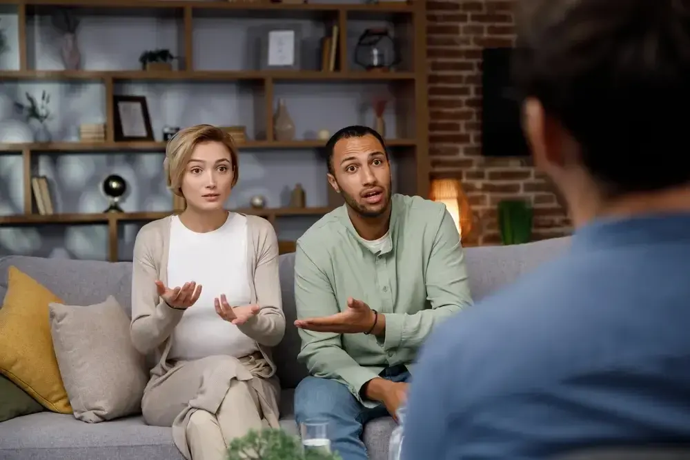 A woman and a man on a couch talking with a therapist, shelves of books behind them.
